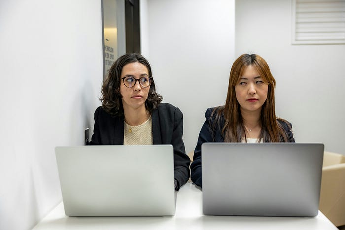 Image 6: two women on laptops side eyeing each other